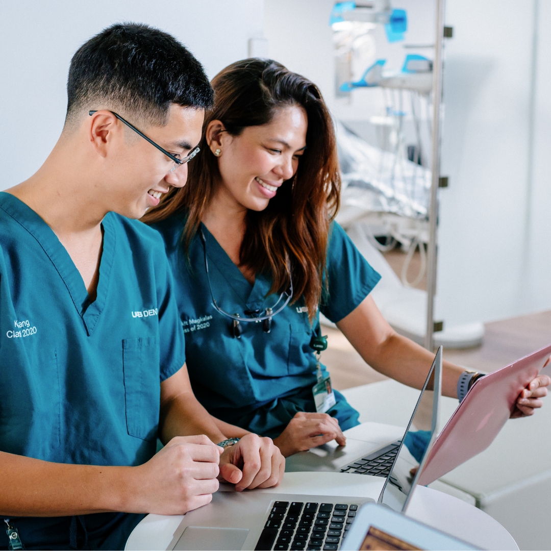 A man and woman in green scrubs sitting in a clinical setting smile at their laptops in their hands